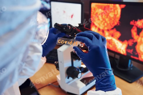 Preview: Scientist in white protective wear indoors in laboratory with lens with COVID-19 title on it