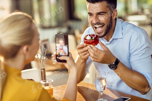 Preview: Happy man being photographed by his girlfriend while eating donut in a cafe,