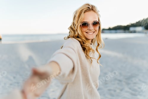 Preview: Curly woman in cashmere sweater smiles and calls to follow her. Portrait of girl in sunglasses on b