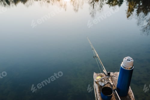 Preview: Fishing rod on a bridge on pond background. Spinning with reel on pier river. Thermos with coffee,
