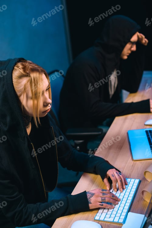 Preview: high angle view of serious female hacker working on new malware with accomplice in dark room