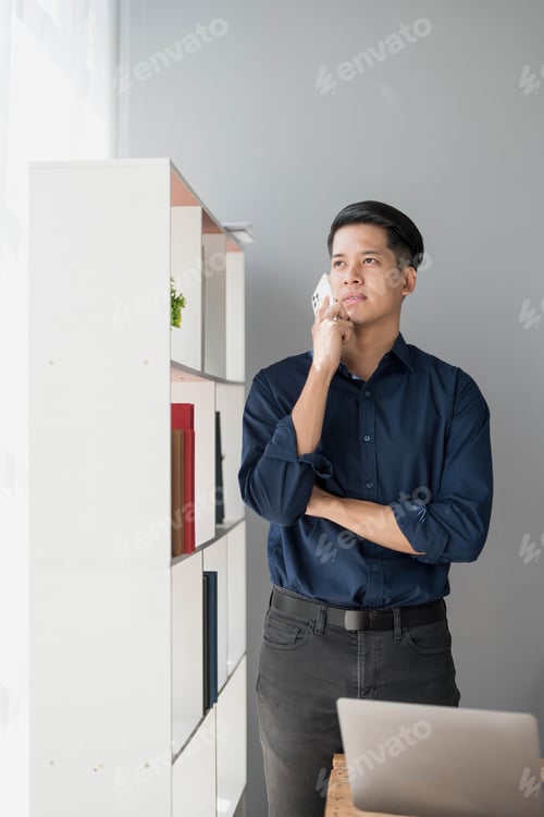 Preview: A young Asian businessman stands on the office of a window talking on the phone.