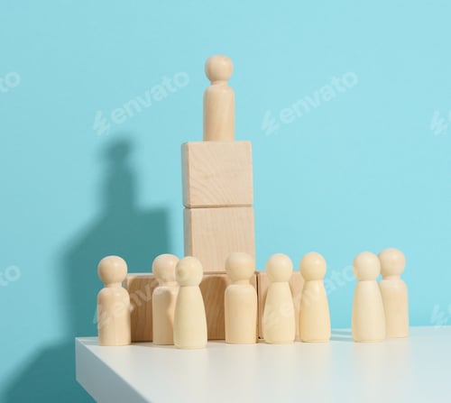 Preview: Wooden figurine of a man stands on a high podium of cubes, below the crowd.