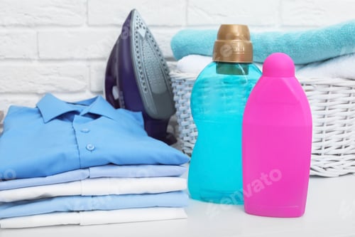 Preview: Stack of clean clothes, iron, detergents and basket with towels on table
