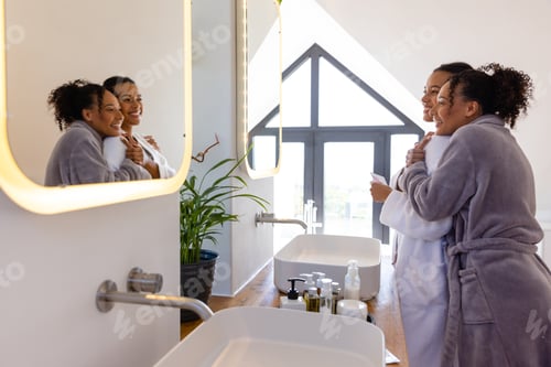 Visualização: Feliz casal de lésbicas birraciais em roupões de banho se abraçando e olhando no espelho no banheiro