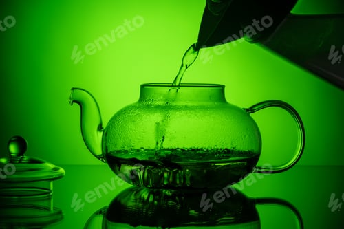 Preview: Pouring hot water into a glass transparent teapot with herbal tea on a green gradient background