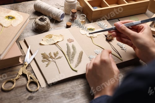 Preview: Woman putting dry flowers into notebook at wooden table, closeup