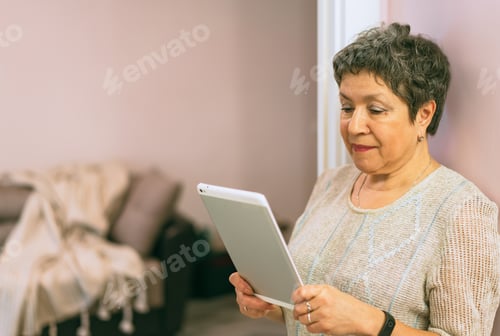 Preview: Attractive elegant senior woman staying near the wall and using a tablet computer at home.