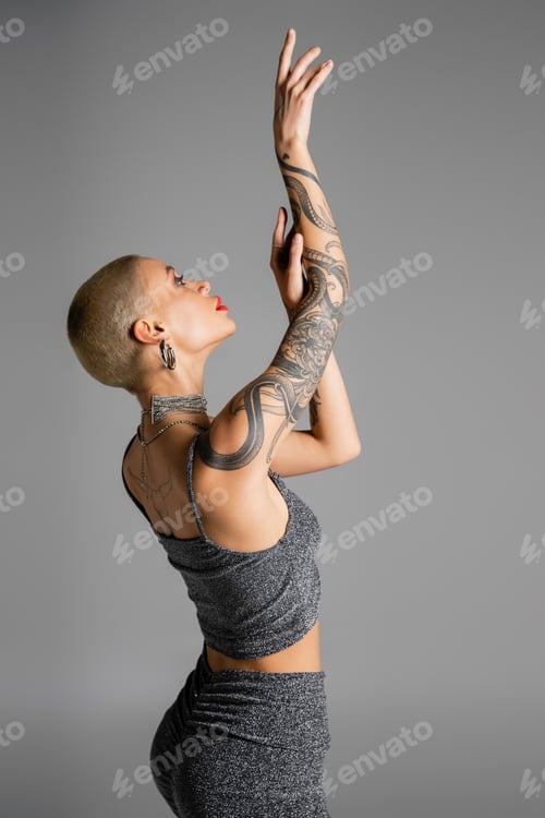 Preview: side view of trendy woman in lurex crop top posing with raised tattooed hands isolated on grey