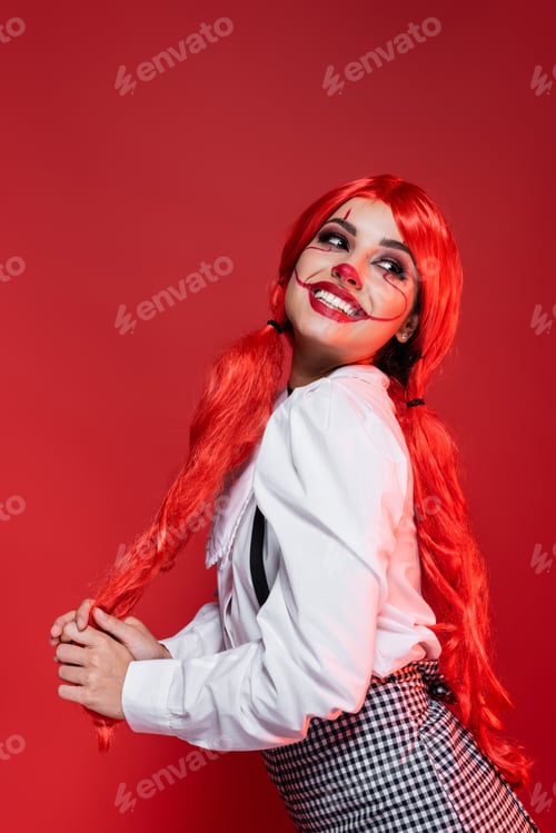 Preview: excited woman with colored hair and clown halloween makeup posing isolated on red