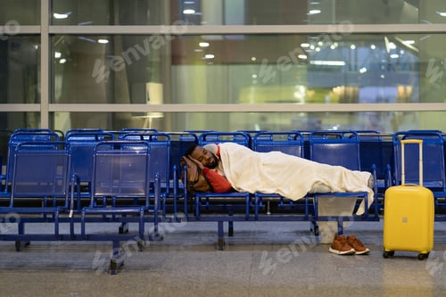 Preview: Young African passenger man sleeping under plaid waiting for flight feel cold. Long night connection