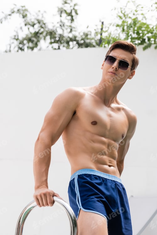 Preview: low angle view of man in swimming trunks and sunglasses looking away outdoors