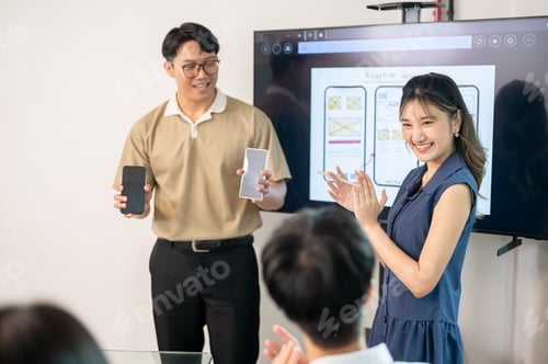 Preview: Asian man standing in front of monitor holding phones presenting app to coworkers in office meeting.