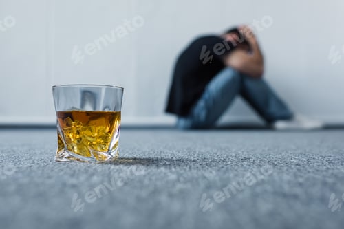 Preview: selective focus of depressed man suffering while sitting on grey floor near glass of whiskey