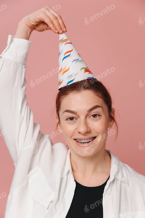 Preview: Portrait of woman smiling with braces and grimacing and holding birthday hat on pink background