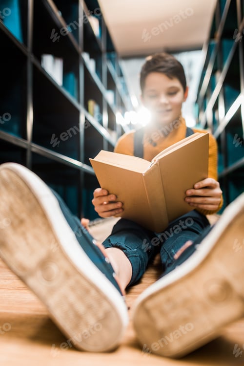 Preview: selective focus of schoolboy reading book and sitting on floor in library