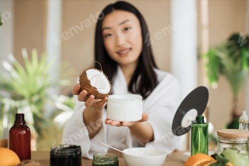 Preview: Focus on coconut and glass jar, young asian woman holding half of coconut and making homemade cream