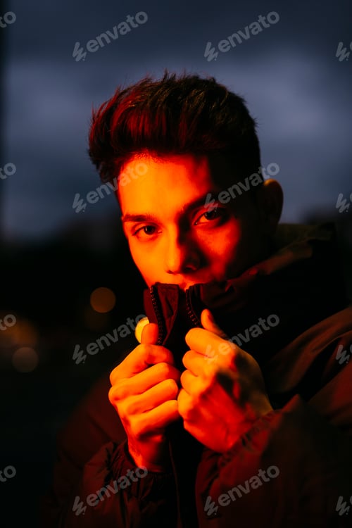 Preview: Young man wearing black puffy jacket pulling up collar under red city lights at dusk