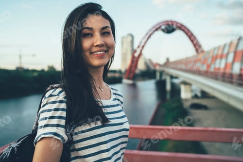 Preview: happy pretty young hipster girl looking at camera standing on river bridge in city
