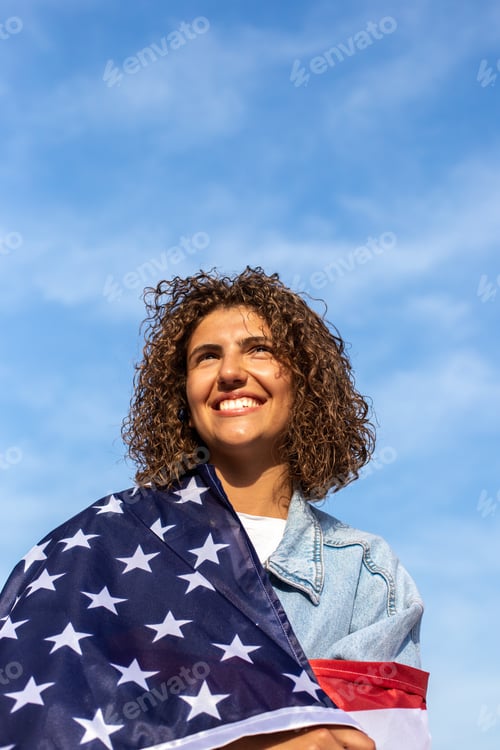 Preview: Happy Beautiful diverse woman holding United States of America flag and standing against blue sky