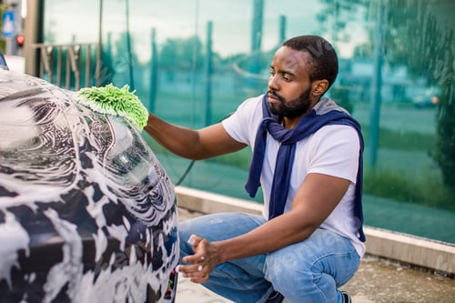 Preview: Young handsome bearded African man in t-shirt and jeans cleaning his electric car