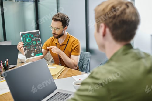 Preview: bearded businessman pointing at infographics near laptops and blurred colleague in modern office