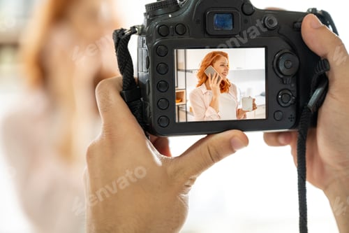 Preview: Closeup of hands holding a professional DSLR camera and taking a shot of red haired woman.