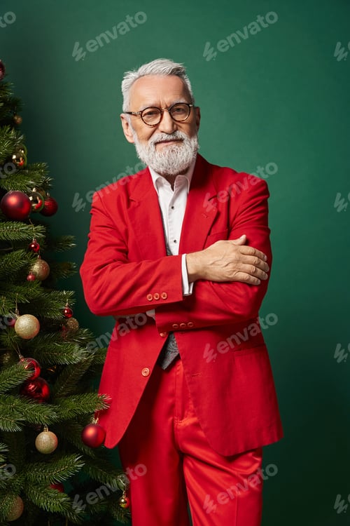 Preview: elegant Santa in red suit with glasses posing near Christmas tree with crossed arms, winter concept