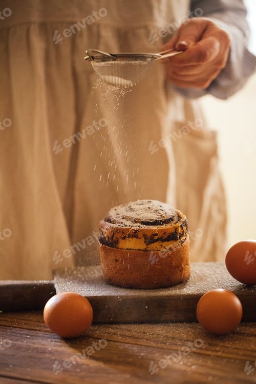 Preview: Person in beige apron sifting powdered sugar over freshly baked cake