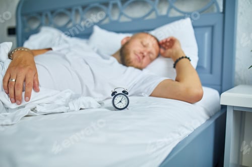 Preview: Bearded man is sleeping alone on a big and cozy bed white linens with alarm clock