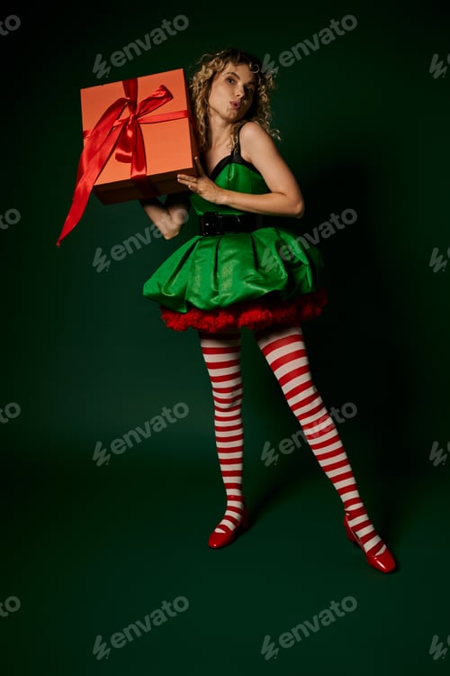 Preview: dreamy new year elf with pouted lips in green dress and striped stockings holding huge gift