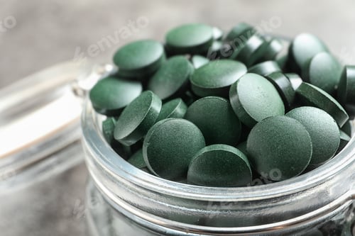 Preview: Glass jar with green spirulina pills on grey table, closeup