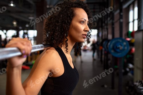 Preview: Young pretty african american sports woman in fitness gym lifting up empty bar in gym, doing warm up
