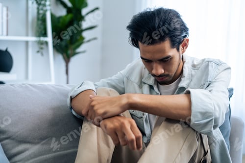 Preview: Asian young upset depressed man sitting alone in living room at home.
