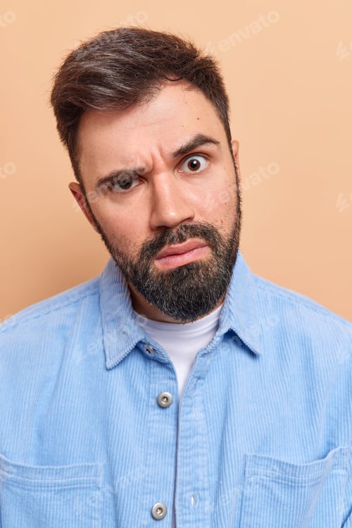 Vista previa: Close up shot of serious unshaven young European man looks attentively and puzzled raises eyebrows f