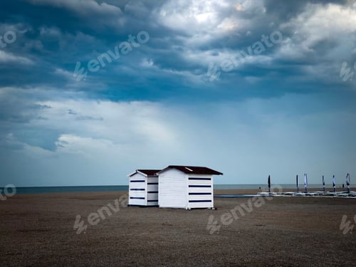 Vista previa: Two small wooden houses on an empty beach in summer and dark storm clouds in the sky