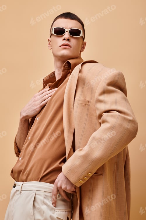 Preview: Portrait of man in beige jacket and shirt with sunglasses and hand near collar on beige backdrop