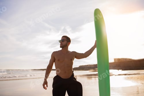 Preview: Young Handsome Male Surfer With Surfboard Standing On Beach At Sunset