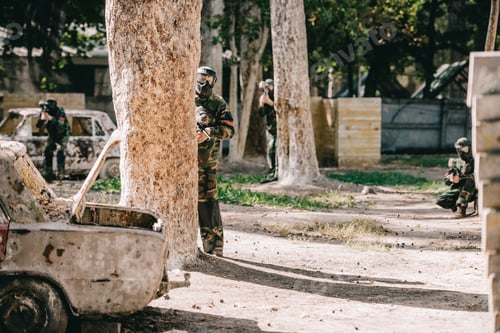 Preview: paintball team in uniform and protective masks playing paintball with marker guns near broken car