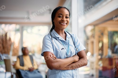 Preview: Happy black nurse with arms crossed in nursing home looking at camera.