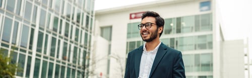 Preview: Smiling indian businessman in suit and glasses with laptop near office building