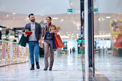Preview: Young happy couple with gift boxes and shopping bags walking through the mall.