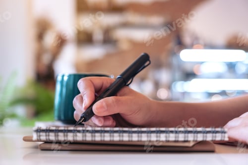 Preview: Closeup image of a woman's hand writing on blank notebook with coffee cup on table in cafe