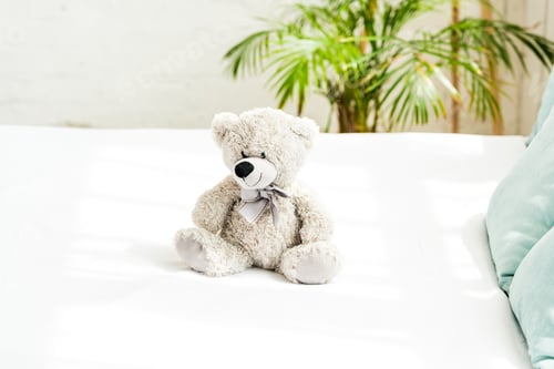 Preview: grey teddy bear near blue pillows on white bedding at home