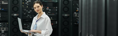Preview: Focused woman in a white shirt works with a laptop in a high-tech server room, banner