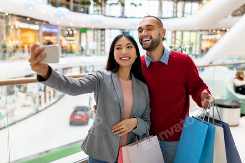 Preview: Happy multinational couple holding gift bags, taking selfie at supermarket, going shopping together