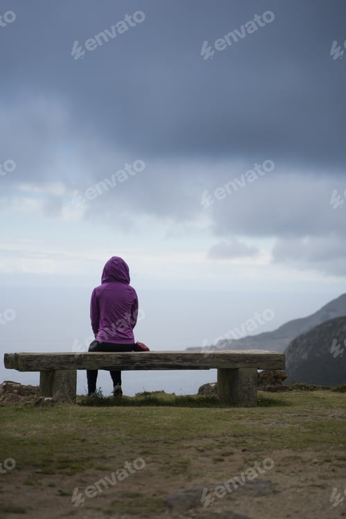 Preview: Thoughtful Hooded person is Looking at Ocean siting on a stone bench