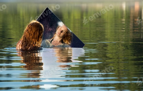 Preview: Young woman standing in lake, holding mirror, looking at reflection