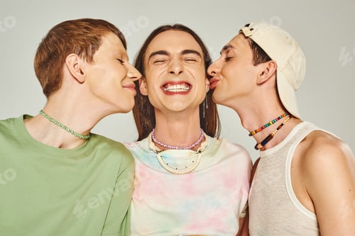Preview: portrait of happy gay men with colorful beads smiling while kissing cheeks of friend on grey