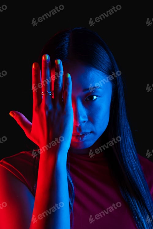 Preview: an asian woman covers Half Face with Hand Looking at the Camera illuminated by red and blue light